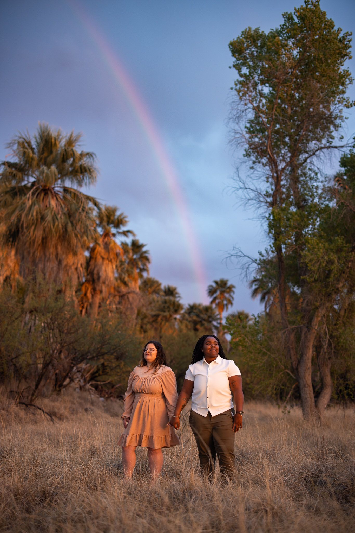 Two people stand together in a desert landscape with palm trees and a vibrant rainbow in the warm evening sky.