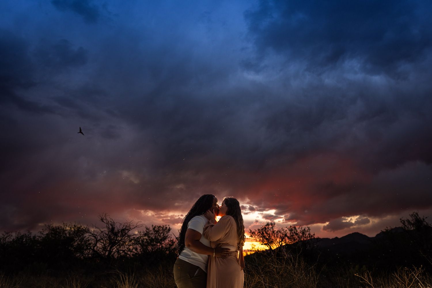 Two silhouettes embrace against a dramatic sunset with dark stormy clouds and mountain backdrop.