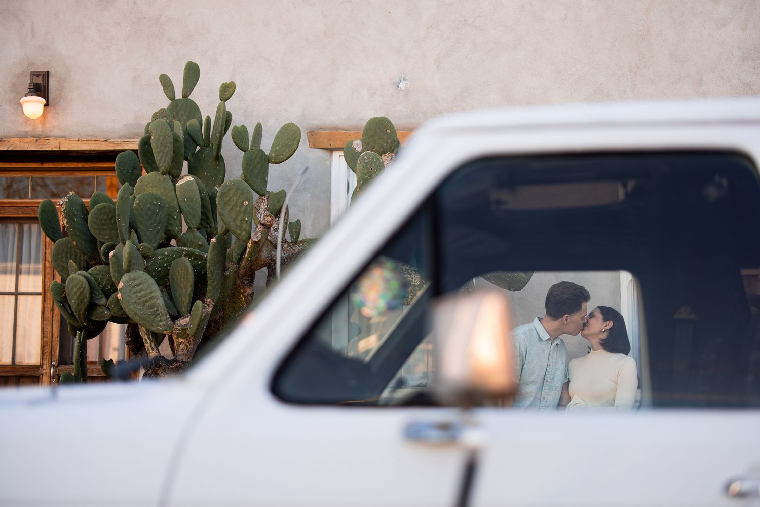 A white vehicle parked next to a large cactus plant growing against a stucco wall with a mounted light fixture.