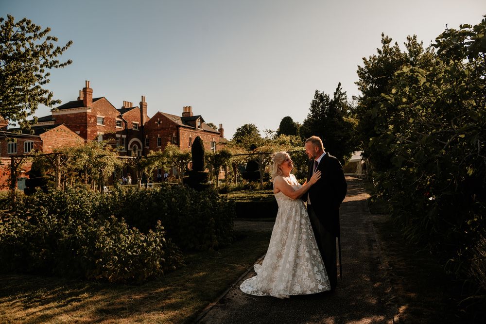Couple shares a romantic sunset moment in a garden setting with a brick building in the background.