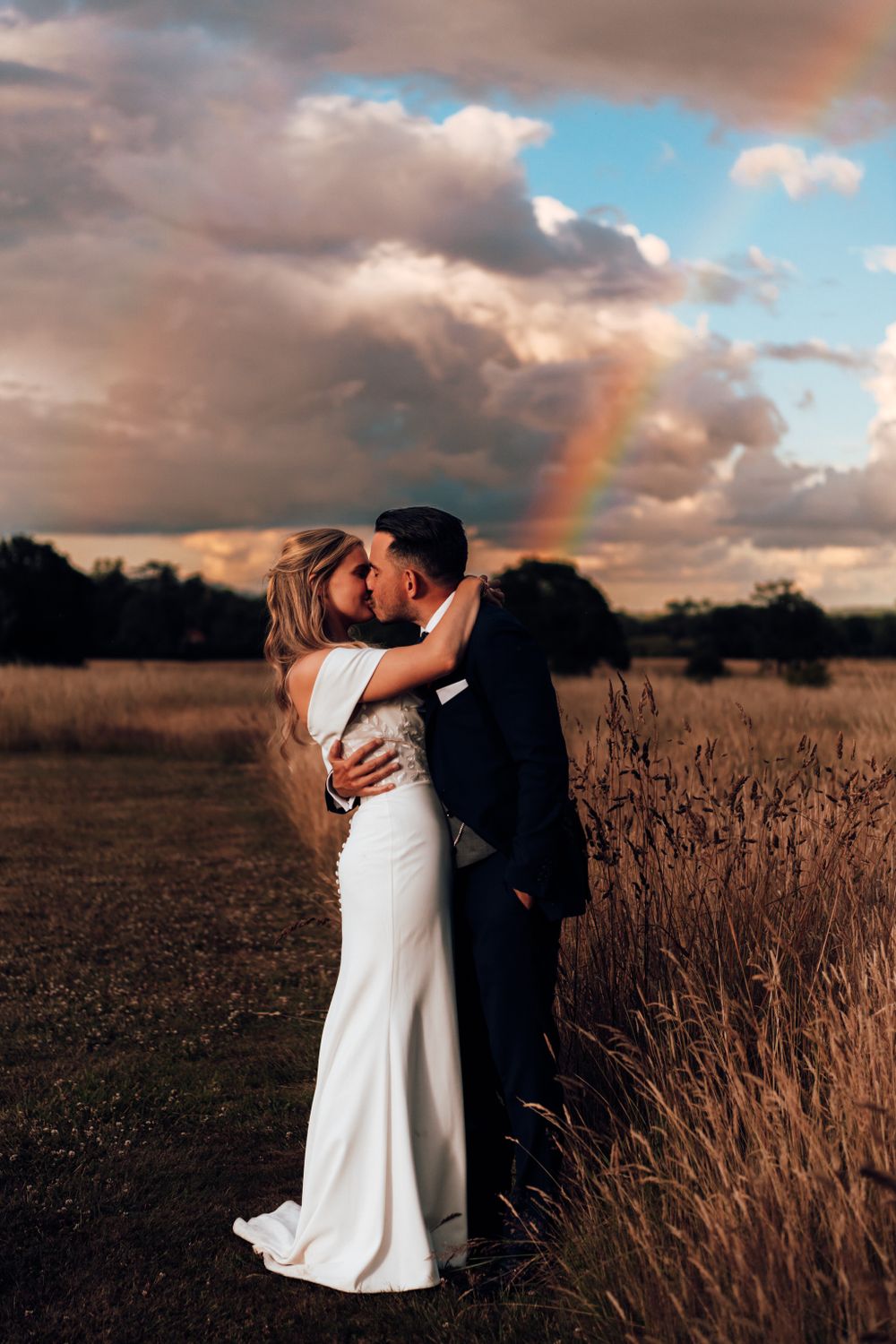 Couple sharing romantic moment in wheat field at sunset with dramatic clouds and rainbow in background.