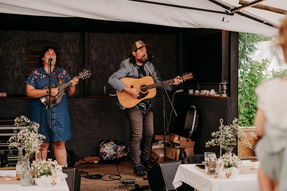 Musicians perform with acoustic guitars under a tent during a wedding reception with decorated tables.