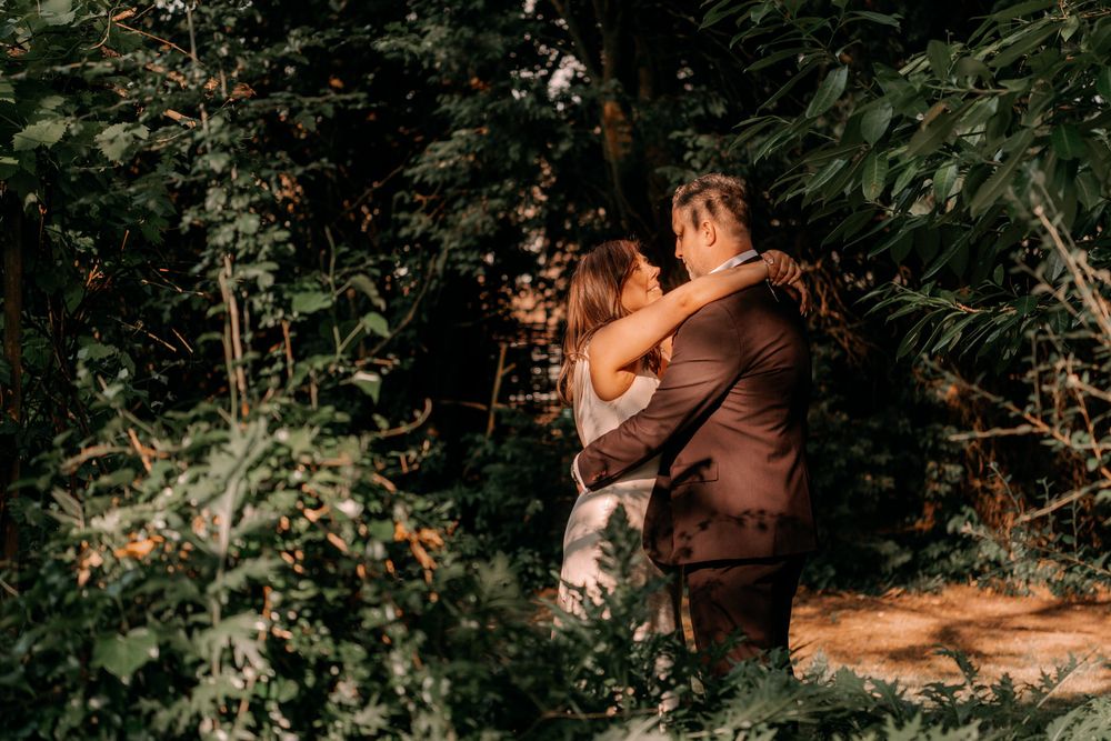 A couple embraces while standing among lush green foliage on a sun-dappled forest path.