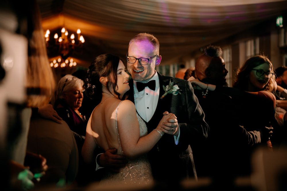 Couple dances intimately at an elegant wedding reception with warm lighting and chandeliers in the background.