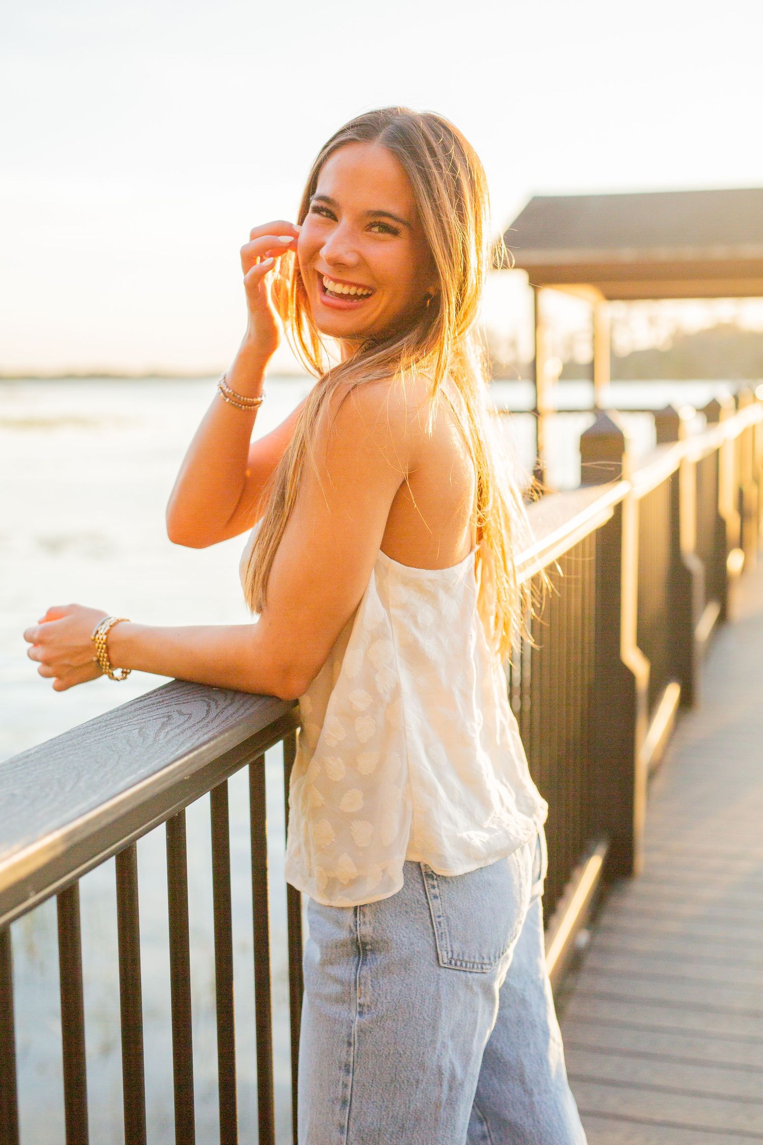 Person standing on wooden pier at golden hour wearing white tank top and jeans while smiling and looking over railing.