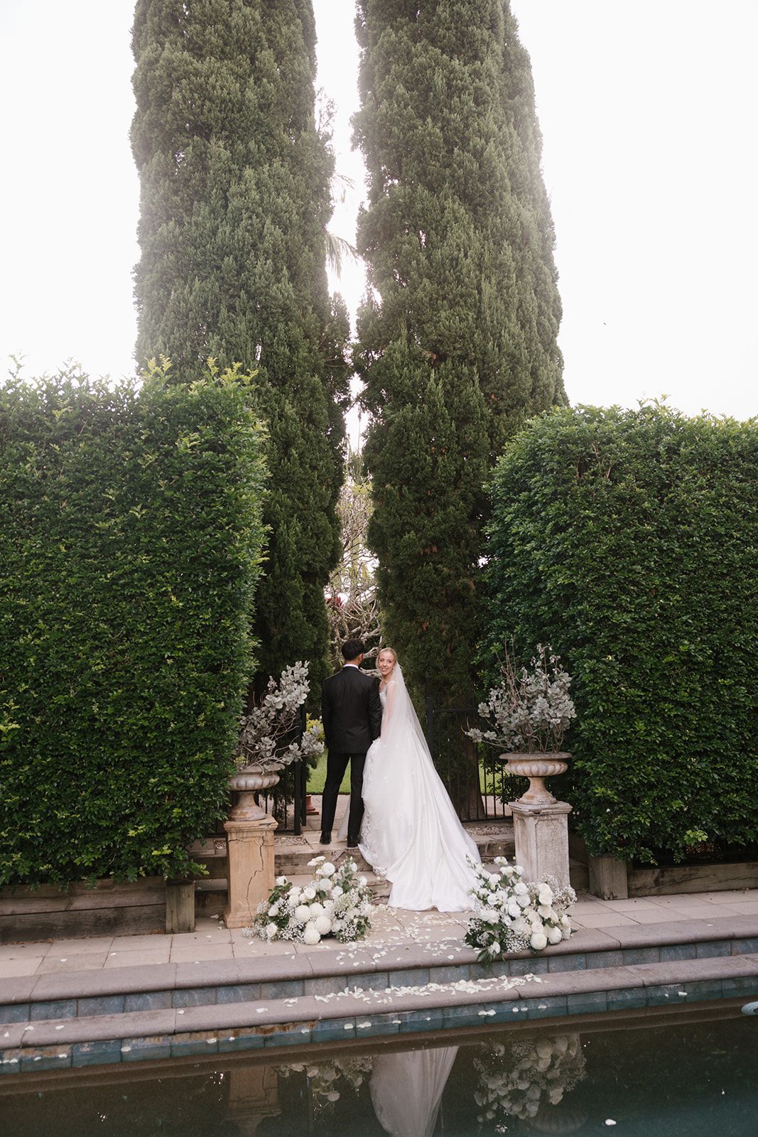 Wedding couple standing between tall cypress trees next to a reflective pool with floral decor.