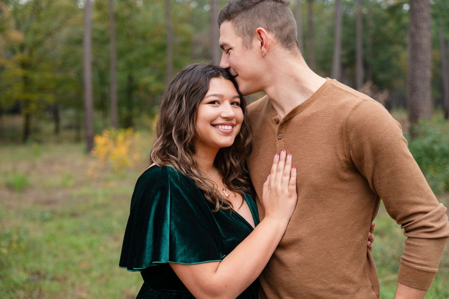 A couple shares a tender moment together in a forest setting wearing a brown sweater and green velvet dress.