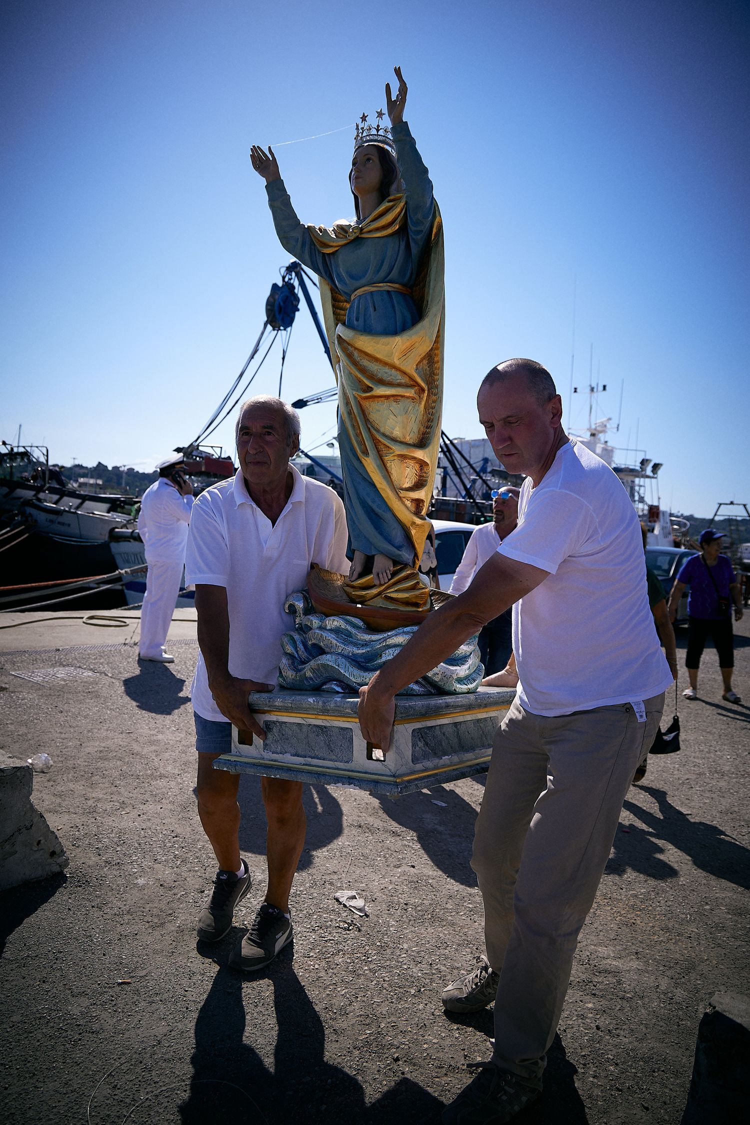 Two people carrying a religious statue on a dock with boats in the background.