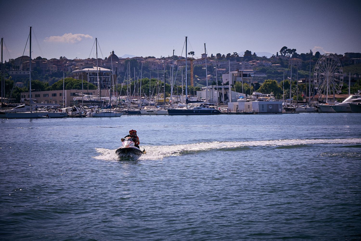 Jet ski rider creates wake across marina harbor with boats and hills in background.
