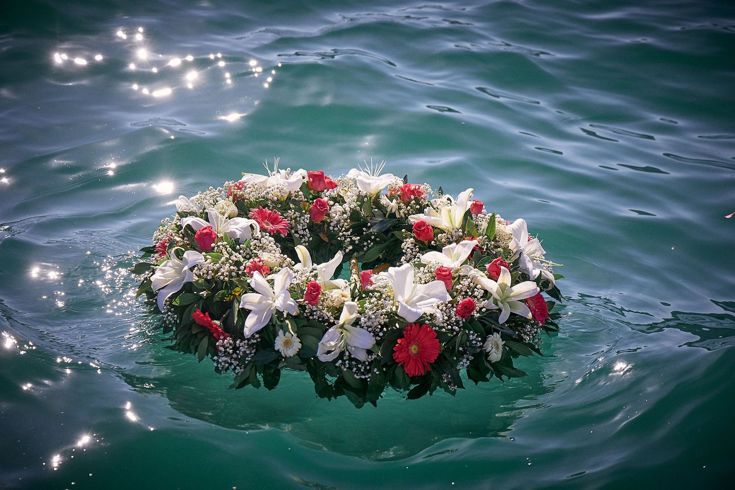 Floral wreath with red and white flowers floating on sparkling ocean water.