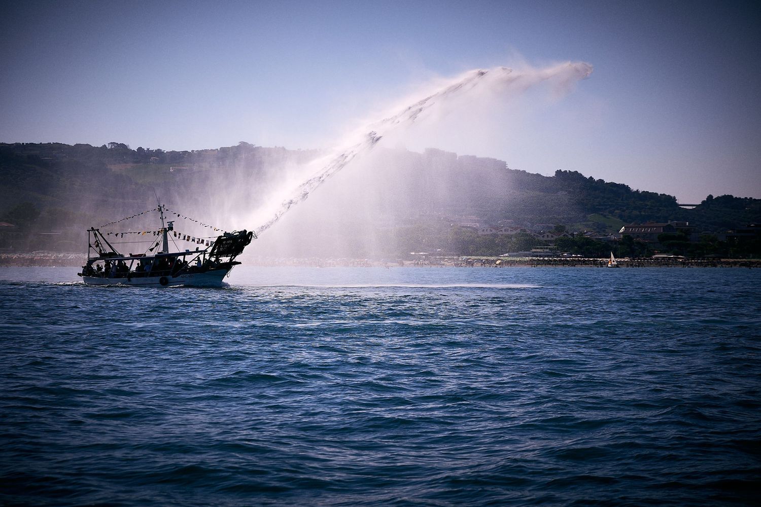 Silhouetted fishing boat sprays water from its side in front of coastal hillside.