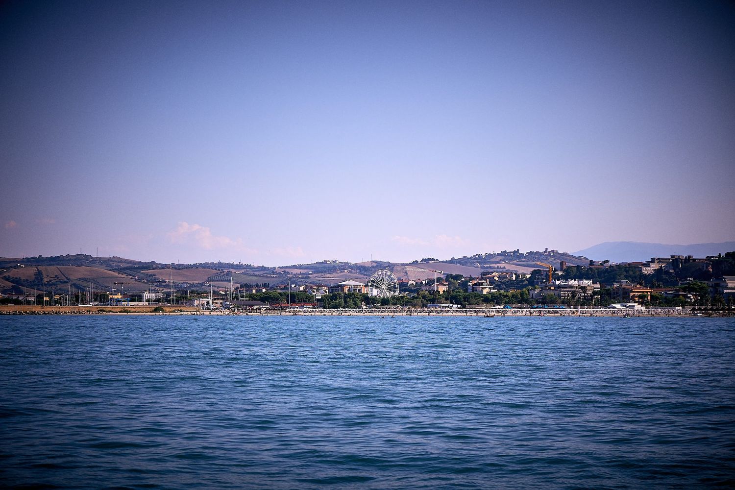 Coastal cityscape view with mountains and buildings along shoreline under clear blue sky.