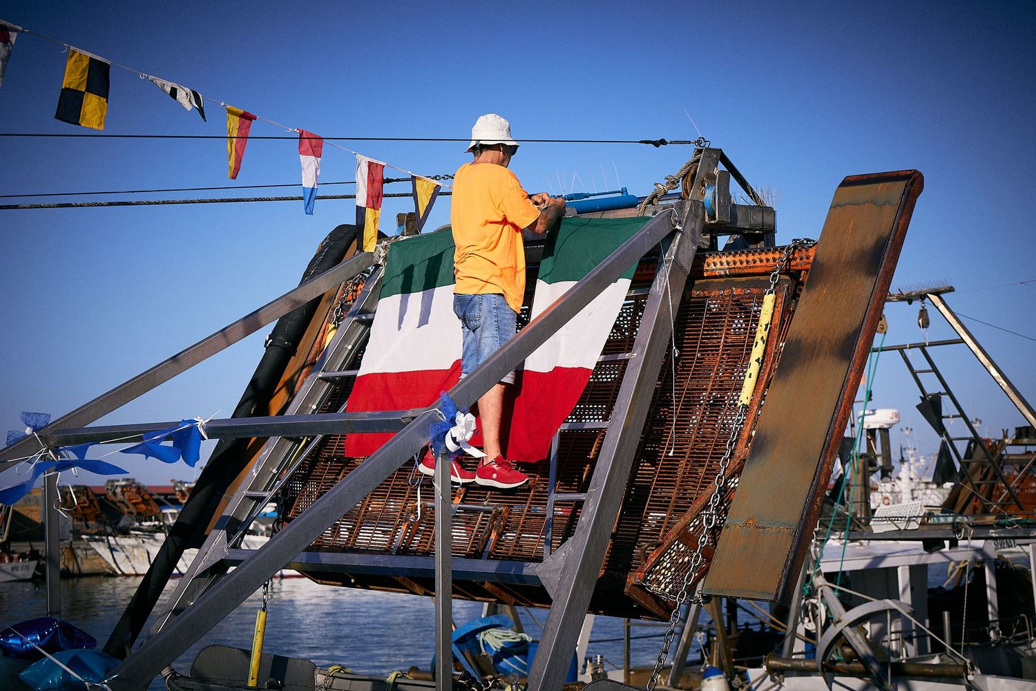 Worker in orange safety vest performs maintenance on equipment with flags visible against blue sky background.