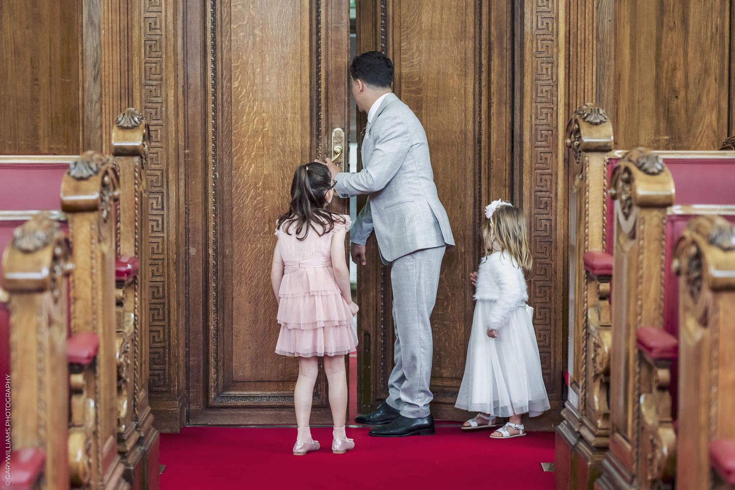 Young children in formal attire stand at ornate wooden doors in a historic building.