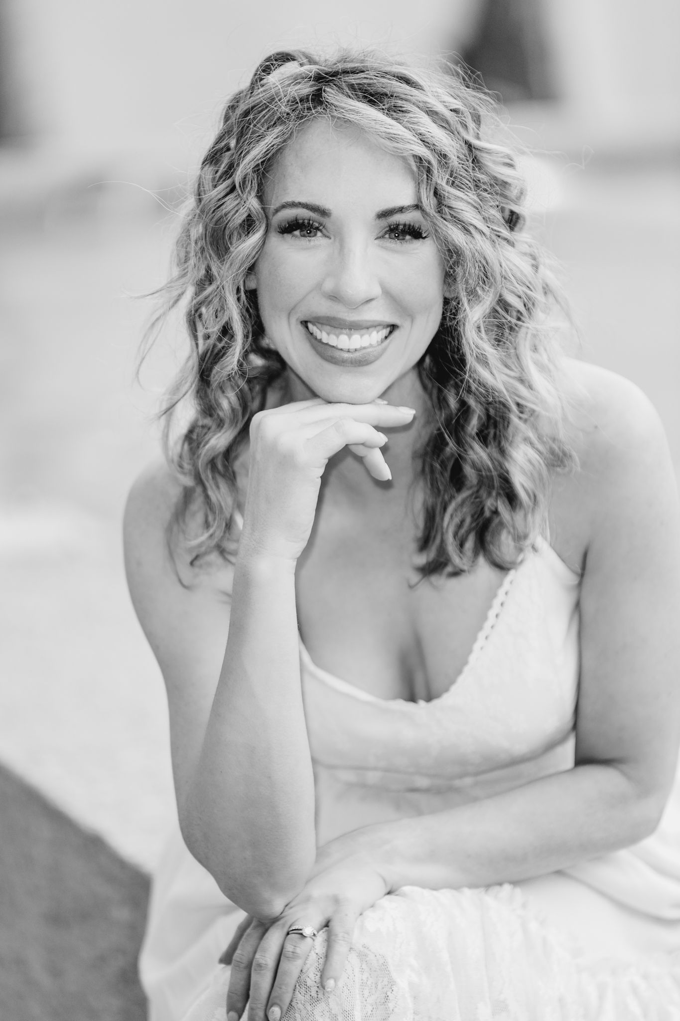 Black and white portrait of a smiling person with curly hair and white dress resting chin on hand.