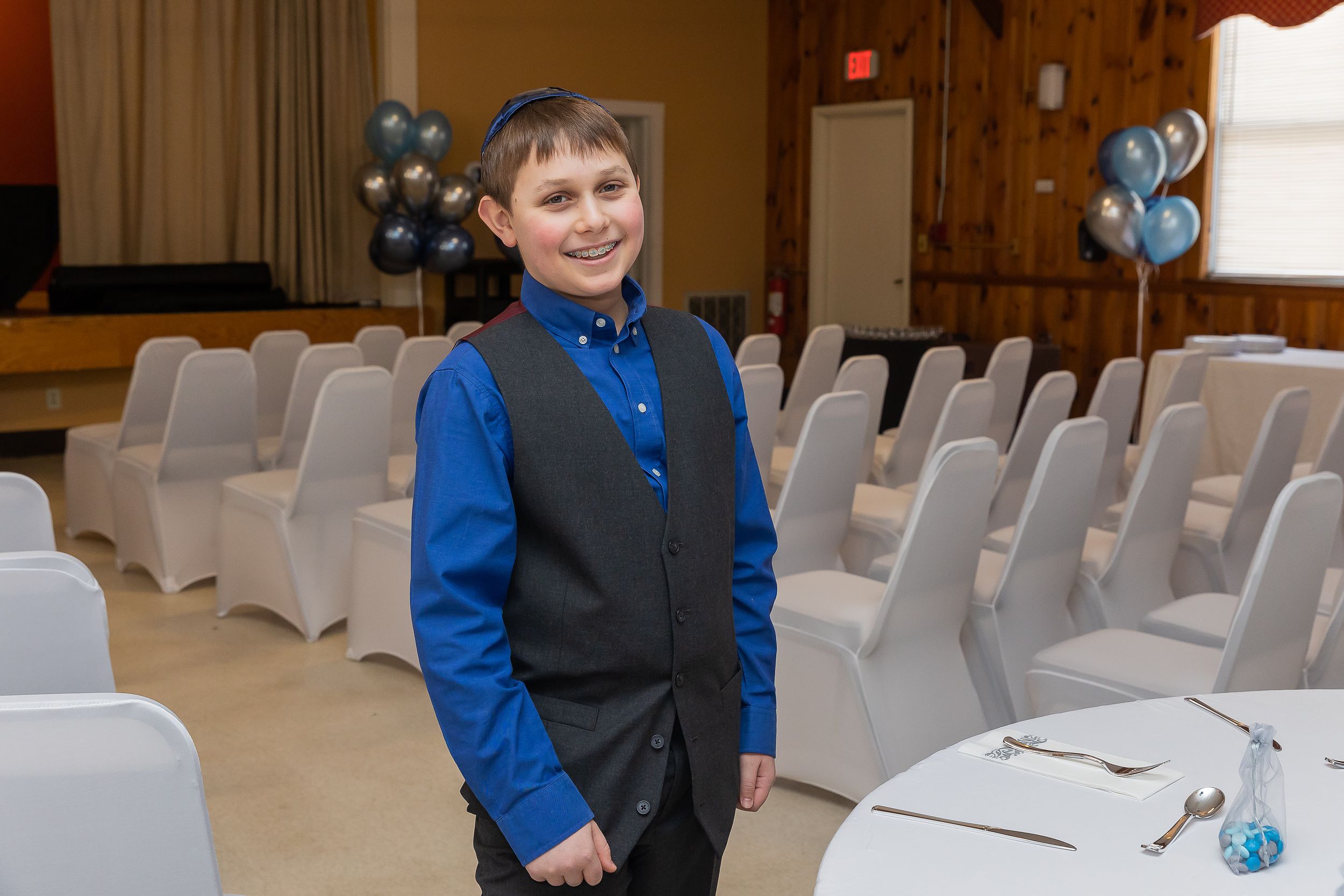 Young person in formal attire standing in event space with white chairs and blue balloons.