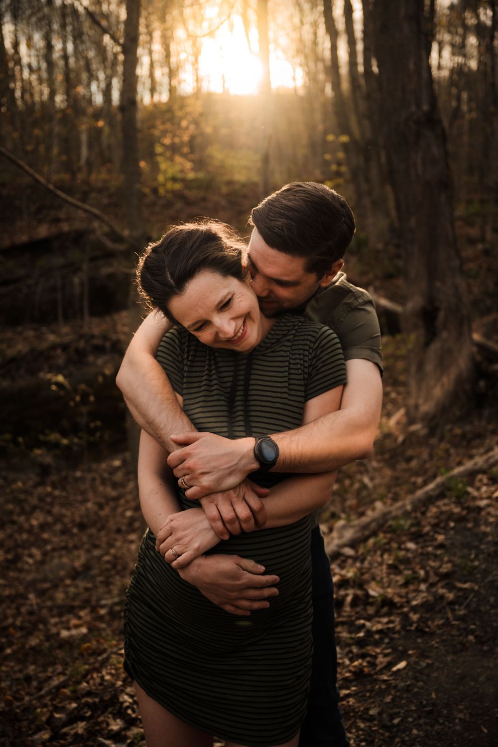 Romantic maternity photo of couple embracing in sunlit forest.
