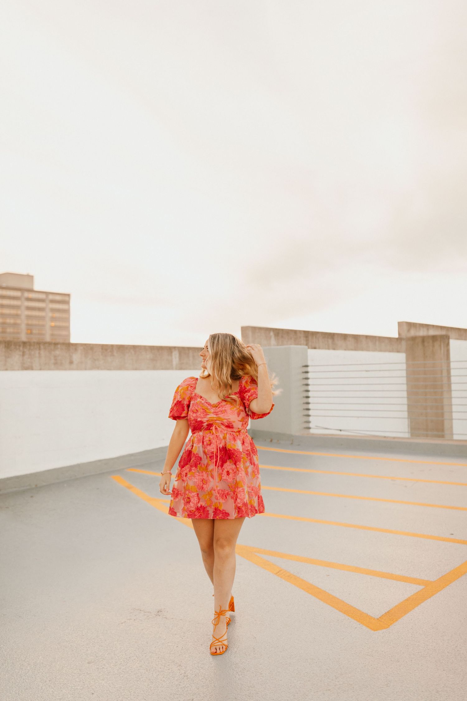 Woman in floral red mini dress walking on empty parking garage rooftop with yellow lines during sunset in Lexington, Kentucky.