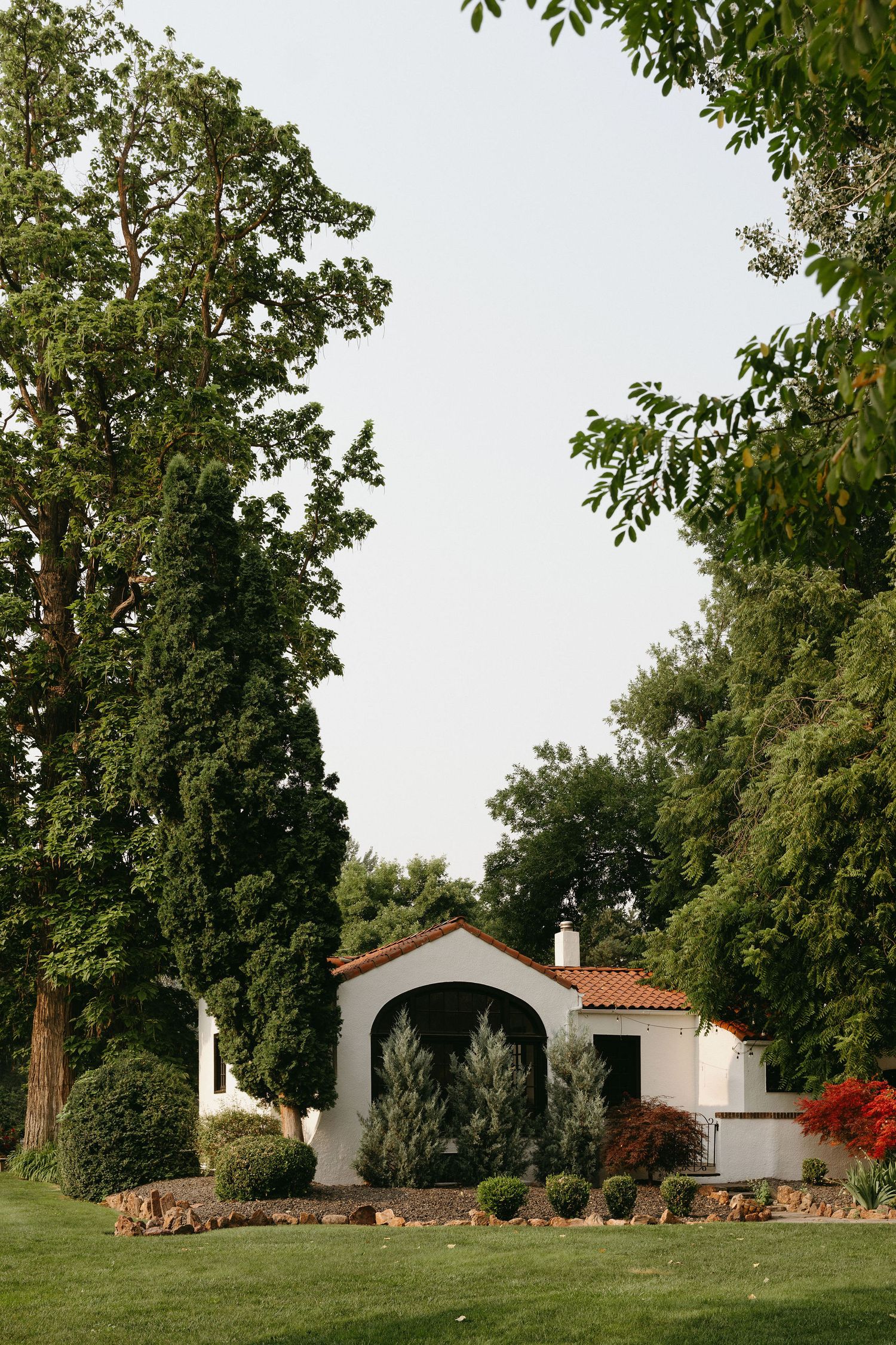 A quaint Spanish-style white stucco cottage with terracotta roof sits nestled among mature trees on a manicured lawn.