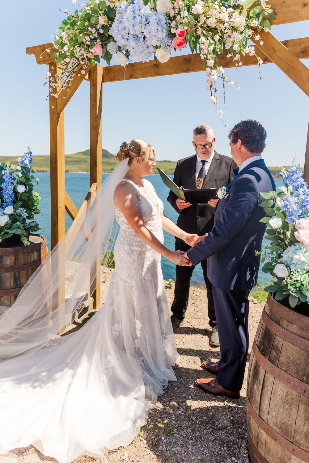 An outdoor wedding ceremony taking place under a wooden arbor decorated with white and blue flowers near the waterfront.