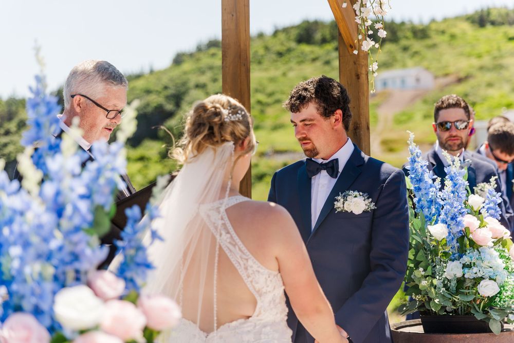 Outdoor wedding ceremony under wooden pergola with blue delphinium flowers and pink roses decorating the scene.