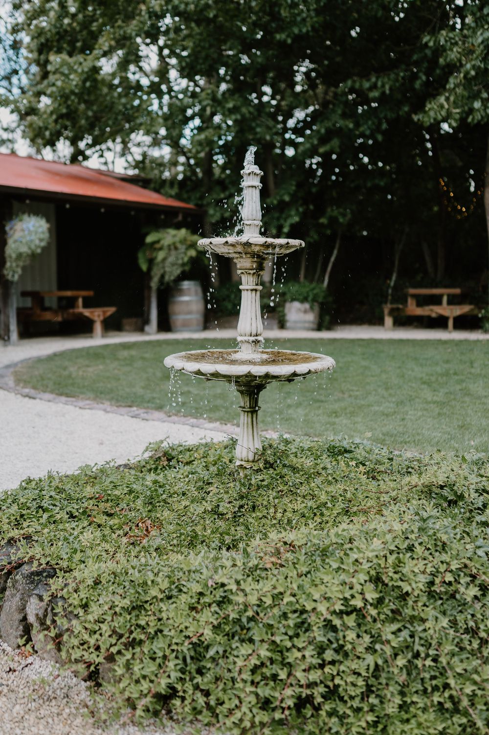 A decorative two-tier stone fountain stands in a peaceful garden setting with benches and greenery in the background.