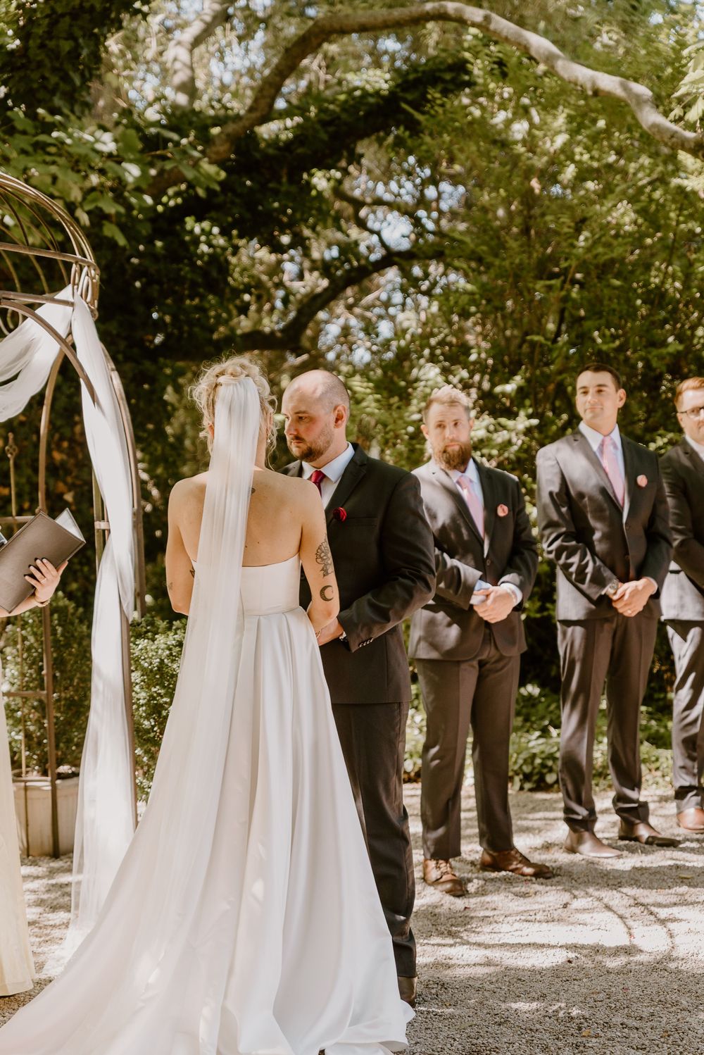 Wedding ceremony under trees with a couple at the altar and groomsmen standing in dark suits on a sunny day.