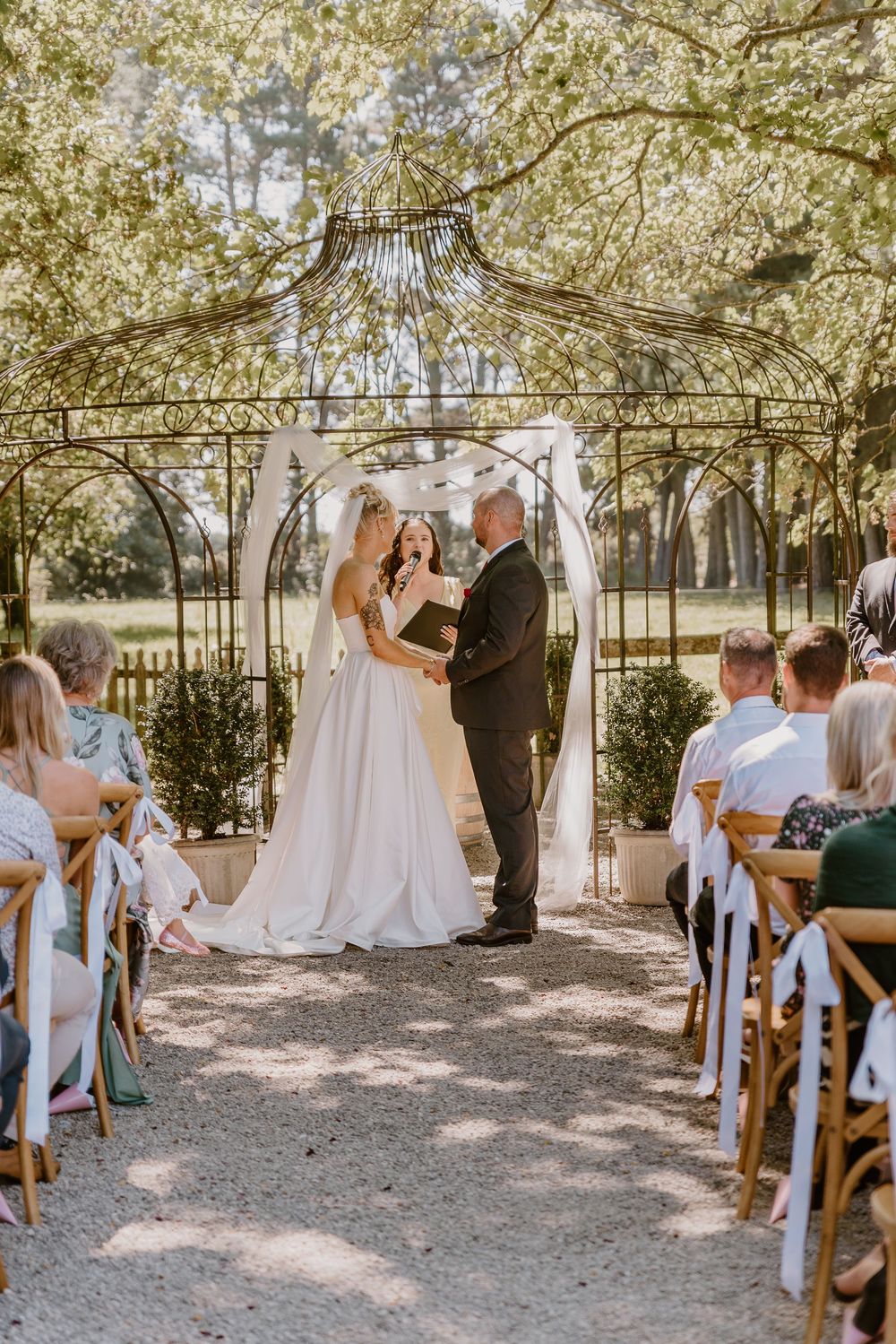 A romantic outdoor wedding ceremony under an ornate iron gazebo with guests seated on wooden chairs in a garden setting.