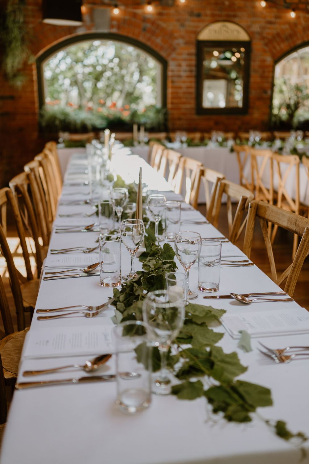 Elegant wedding reception table setting with white linens, natural greenery garlands and rustic wooden chairs in a brick venue.