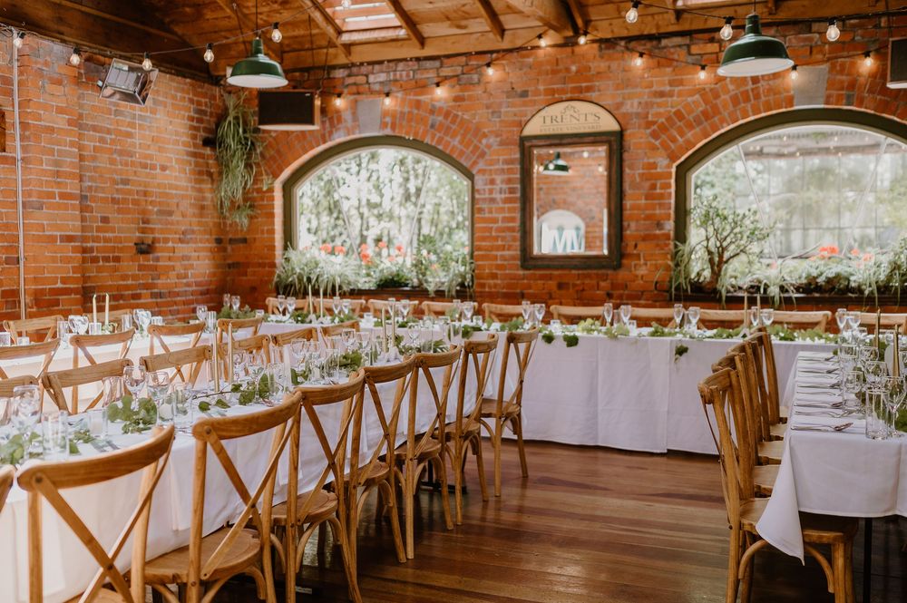 Long rectangular banquet tables with white linens and wooden chairs arranged in a rustic brick-walled reception venue.