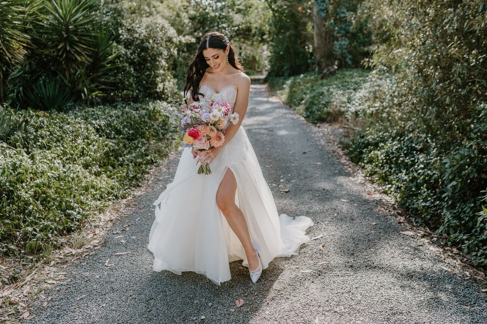 A bride in a flowing white gown with a thigh slit walks down a garden path holding a colorful bouquet.