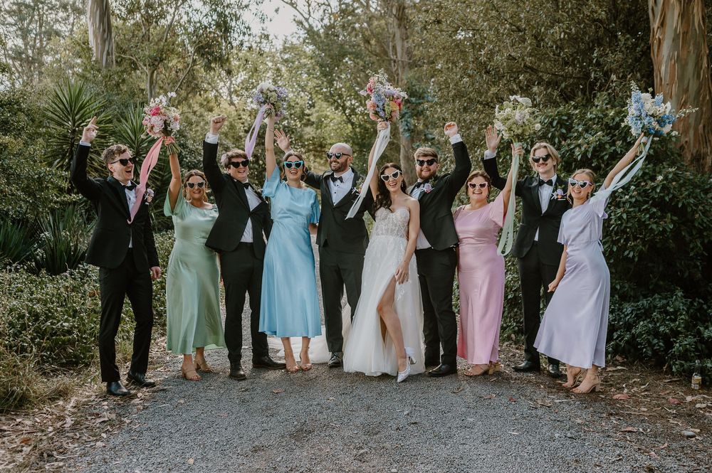 Wedding party celebrating outdoors with raised arms in a garden surrounded by lush greenery and trees.
