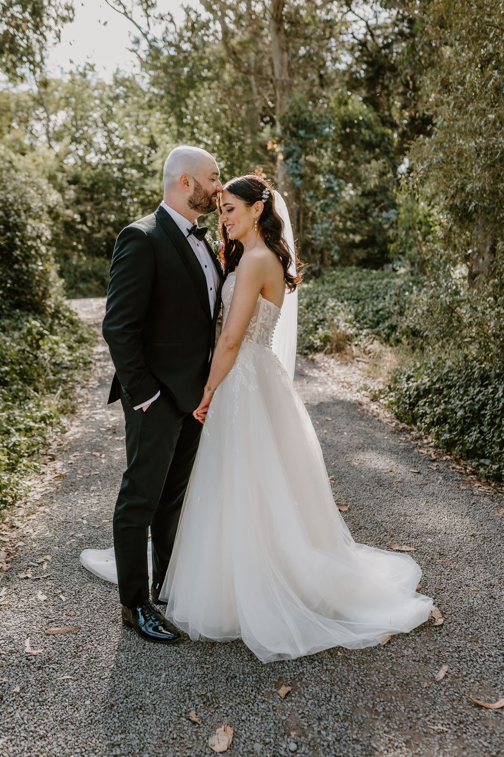 A romantic outdoor wedding moment captured on a tree-lined gravel path showing a couple in formal wedding attire.