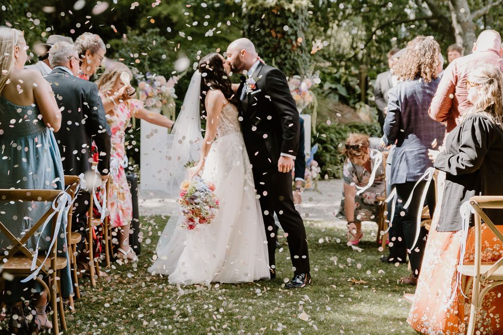 Wedding celebrants toss flower petals and confetti onto festive newlyweds during an outdoor ceremony.