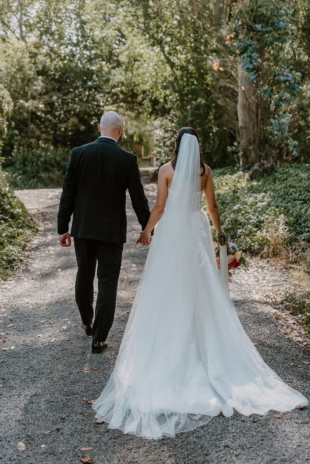 A couple walks down a tree-lined path after their wedding ceremony with the bride's long white veil trailing behind.