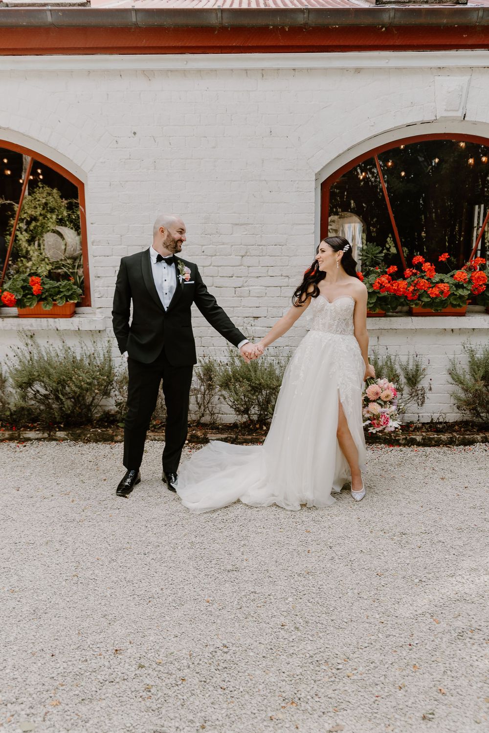A wedding couple playfully walks and spins together on a gravel path next to a white brick wall with red floral accents.
