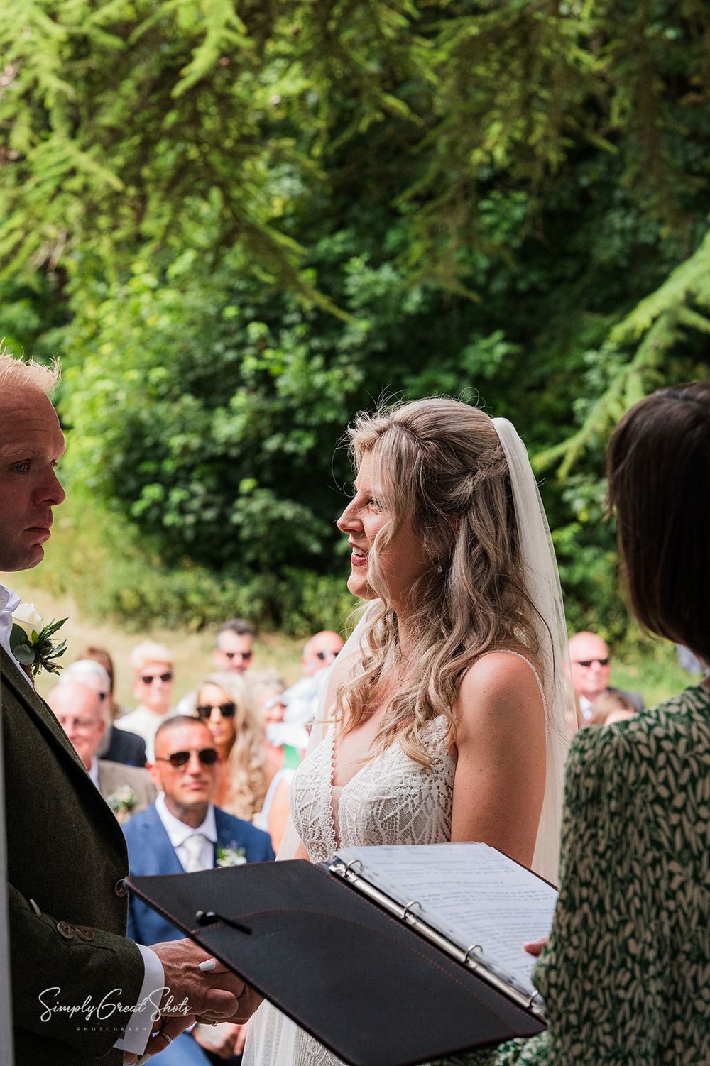 An outdoor wedding ceremony at Fort Henry with the bridal couple standing in front of green trees in summertime, with guests in attendance.