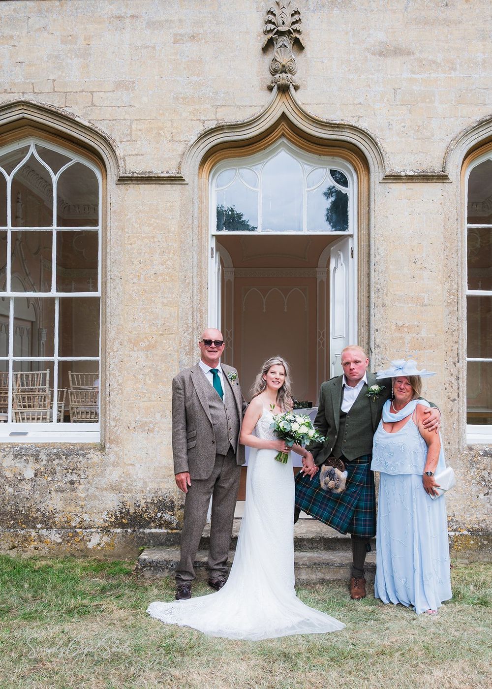 The bride, groom and the bride's parents pose in front of a Gothic revival doorway at Fort Henry wedding venue on a hot sunny day.