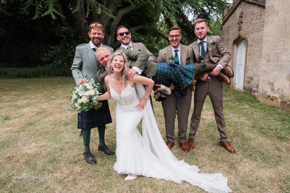 A wedding party in Scottish attire, tweed suits and a white bridal gown pose joyfully on the lawn outside Fort Henry wedding venue.
