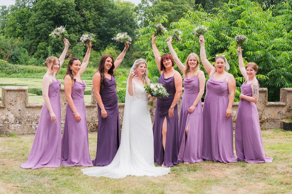 A bridal party poses in flowing purple bridesmaid dresses and a white wedding gown, holding bouquets aloft at Fort Henry Wedding Venue.