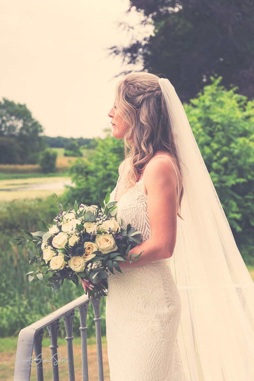 Side profile of a bride in a lace dress holding a white and green floral bouquet looking out towards the lake at Fort Henry Wedding venue.