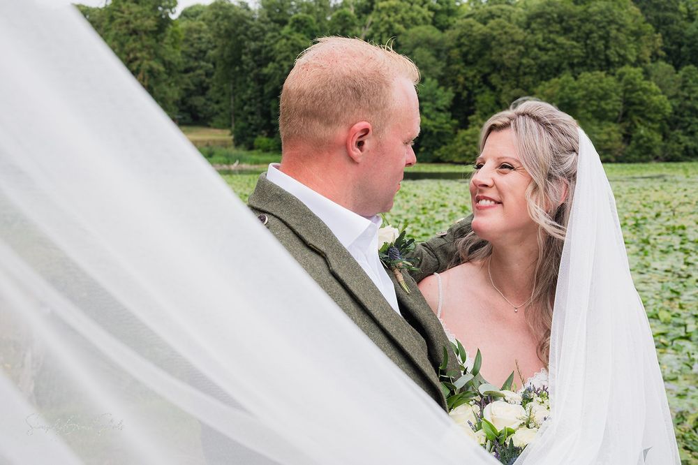 A happy couple shares a joyful moment together on their wedding day with a flowing veil and white and green bouquet.