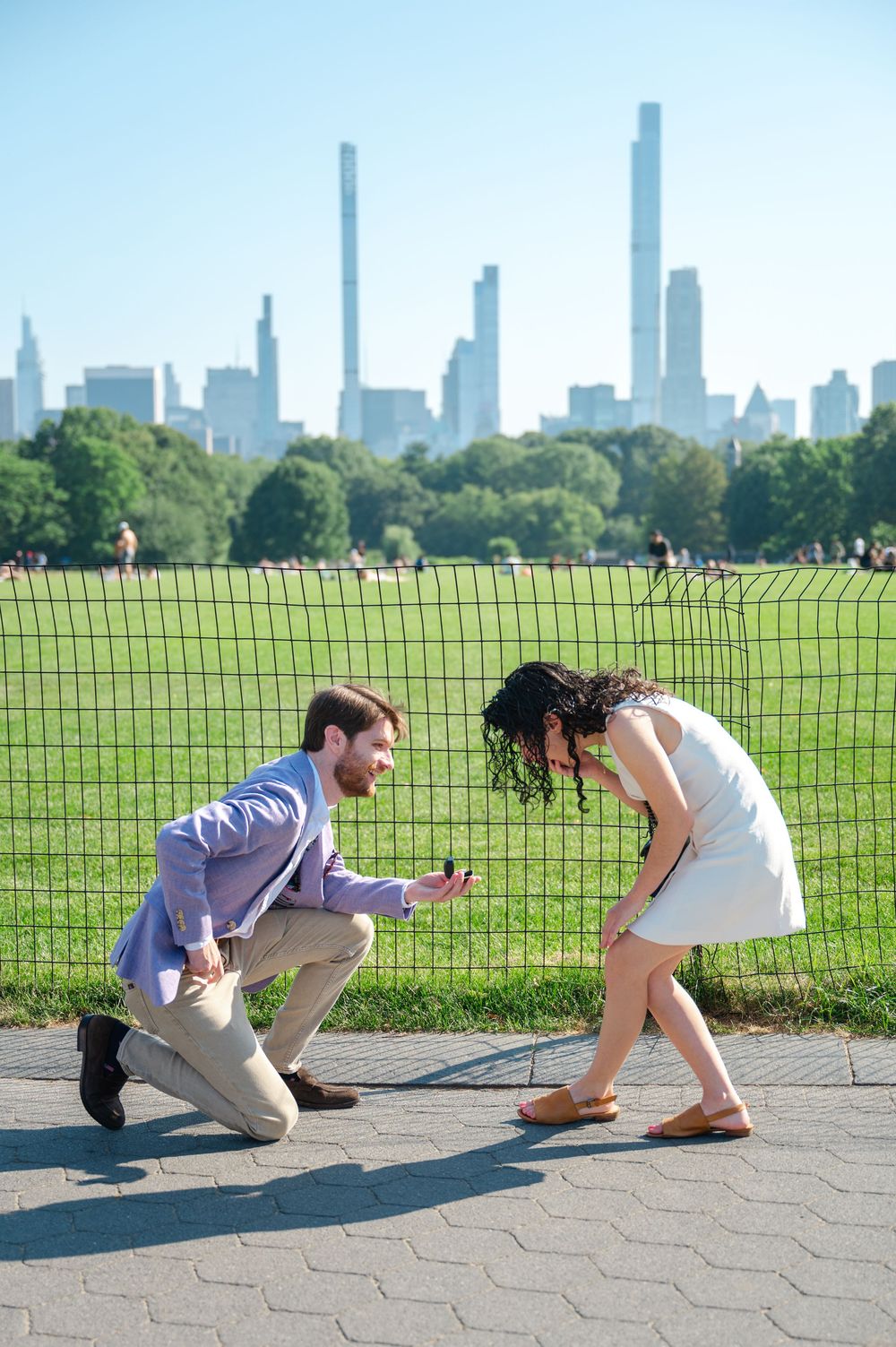 Woman in white bends to see engagement ring during a surprise proposal moment at Central Park with the New York City skyline behind them.