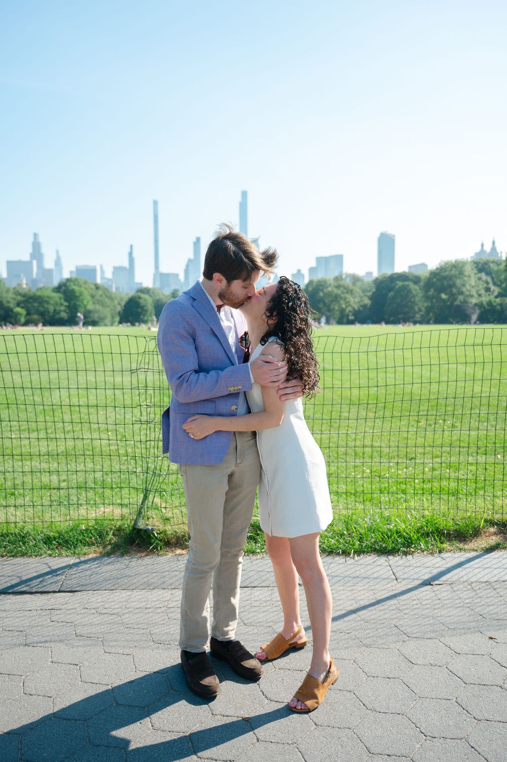 A couple shares a romantic kiss in Central Park with the Manhattan skyline visible in the background. Photo by Joliette Marie Photography