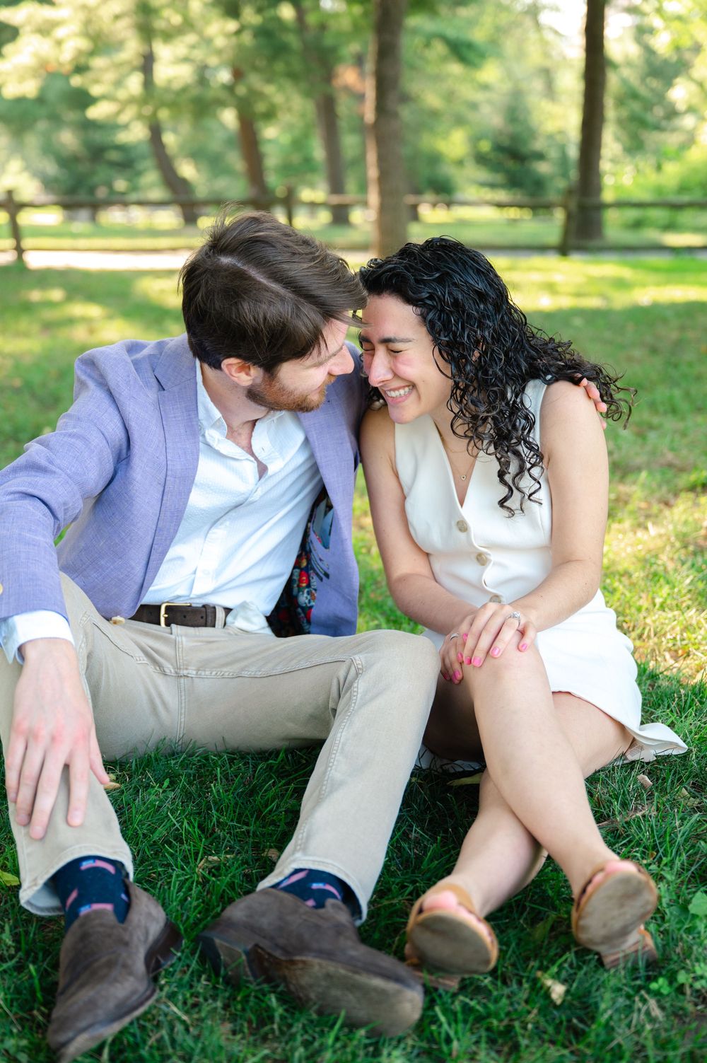 A couple shares an intimate moment while sitting together on green grass in a sunlit park setting.