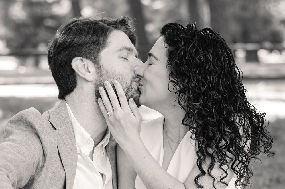 A romantic black and white photo of a couple sharing a tender kiss outdoors with curly hair and formal attire.