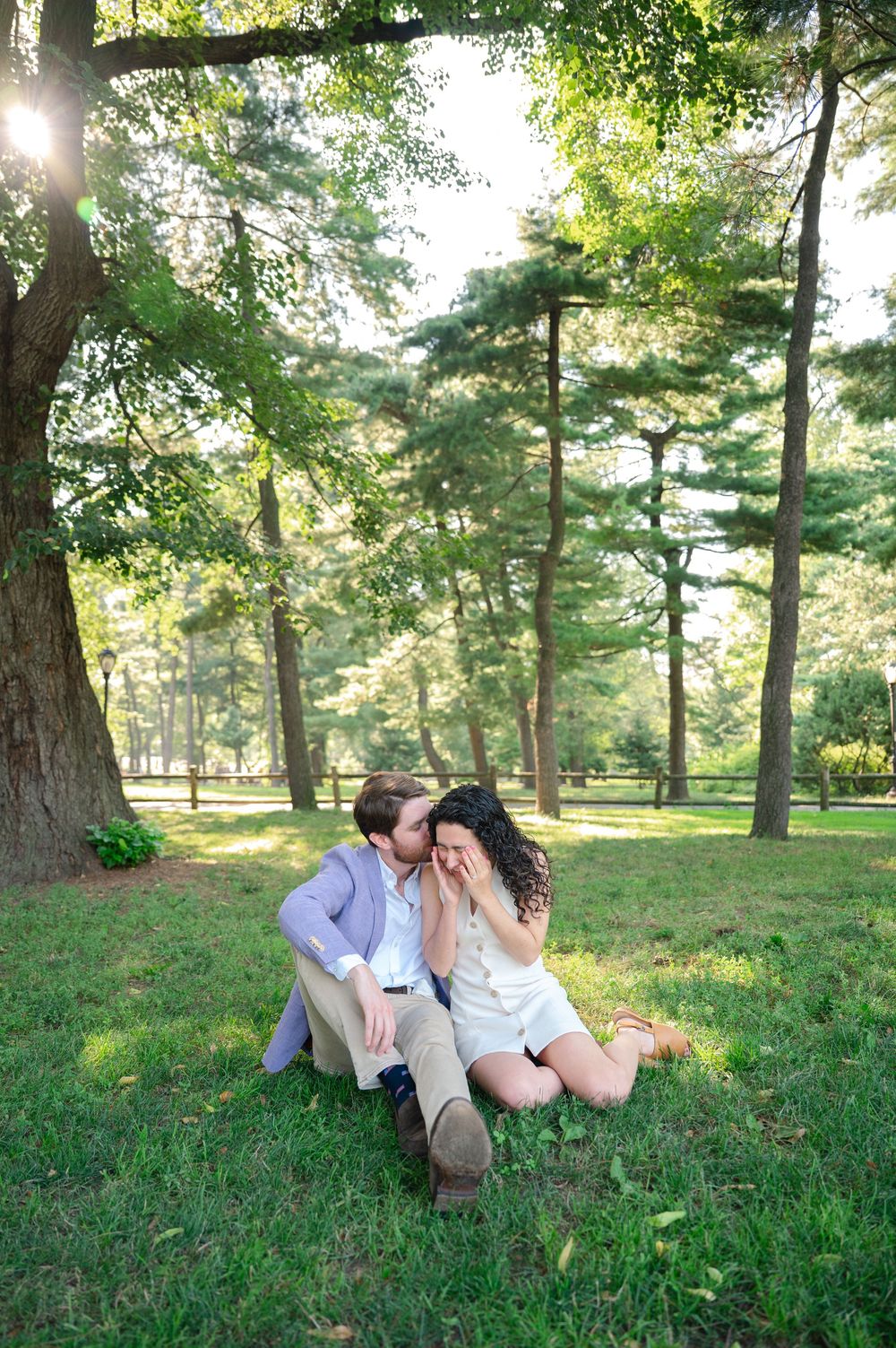 Couple shares intimate moments while relaxing on grass in a sunlit park with tall pine trees during their engagement session.