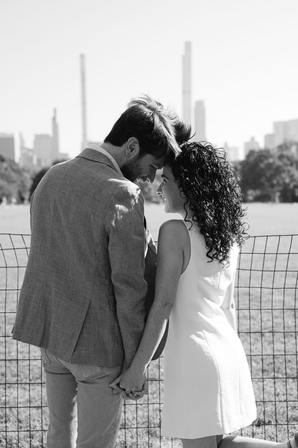 A romantic couple holds hands while leaning together by a wire fence in Central Park, with skyscrapers in the background during a proposal.