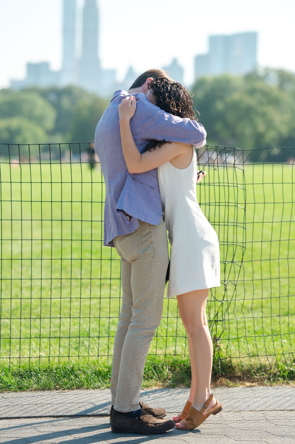 A couple embraces hugs near a fence in Central Park with the Manhattan skyline visible in the background on a sunny day.