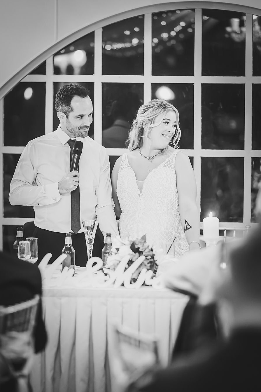 Wedding couple stands at decorated reception table giving speeches in elegant venue with arched windows.